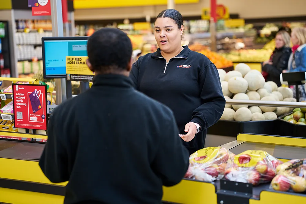 A No Frills cashier is assisting a customer at the checkout counter.