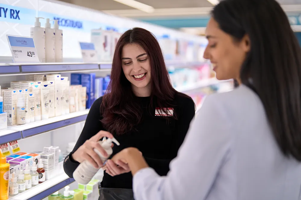 A cosmetics associate is demonstrating a product on a customer's hand in a store aisle