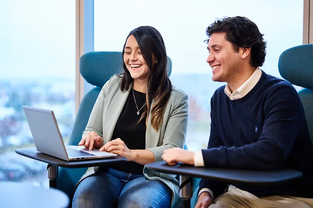 Two young professionals are laughing while working together on a laptop in an office lounge