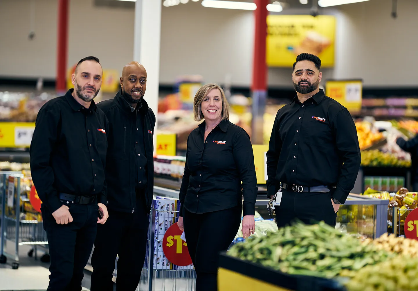 A team of four No Frills employees is posing and smiling in a produce aisle.