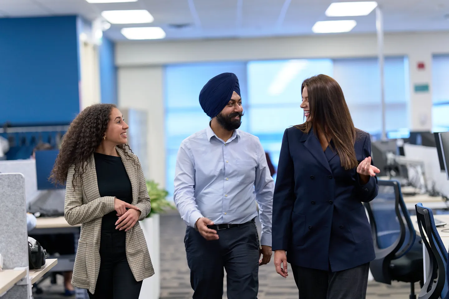 Three diverse and smiling professionals are walking and talking together in an open-plan office