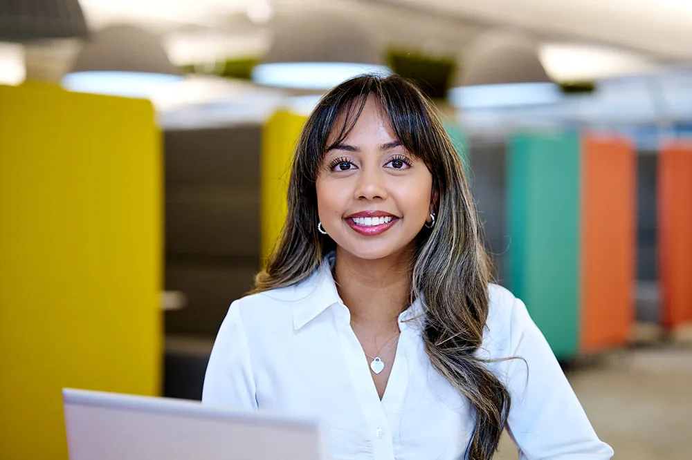 A smiling young professional is sitting in an office space, working on her laptop