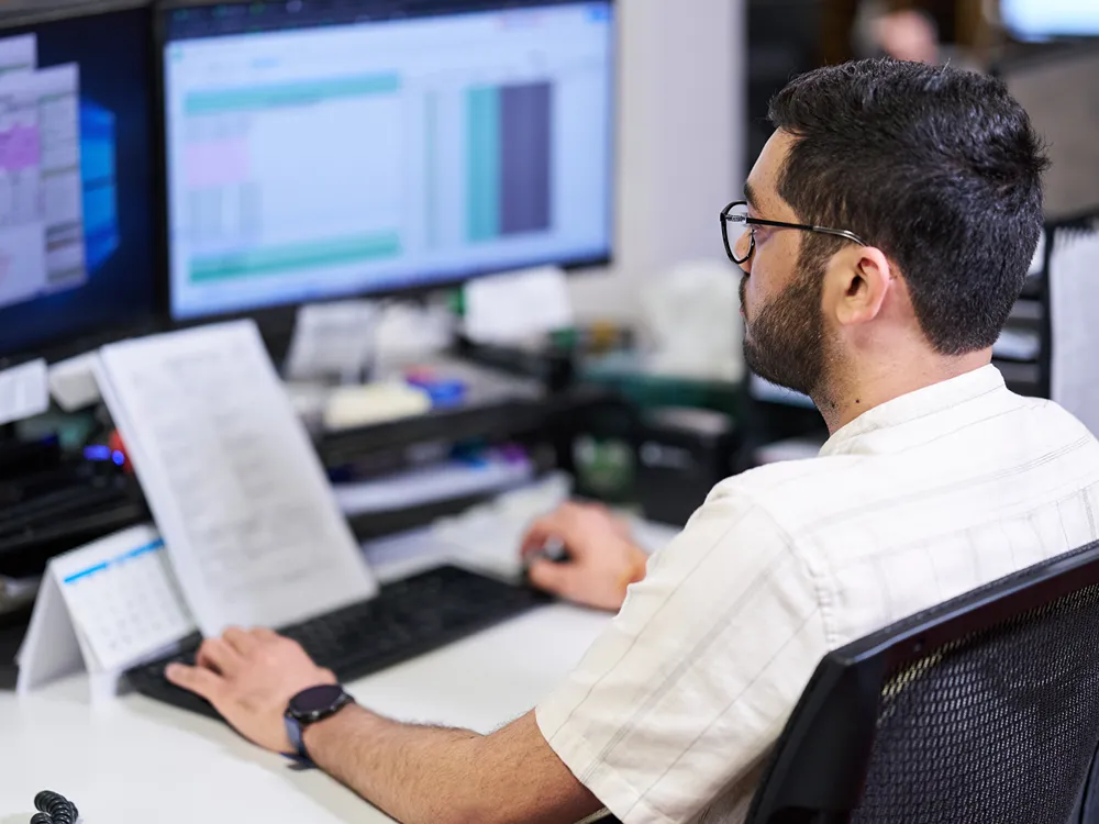 A healthcare systems professional is using two computer monitors to review data at an office desk.