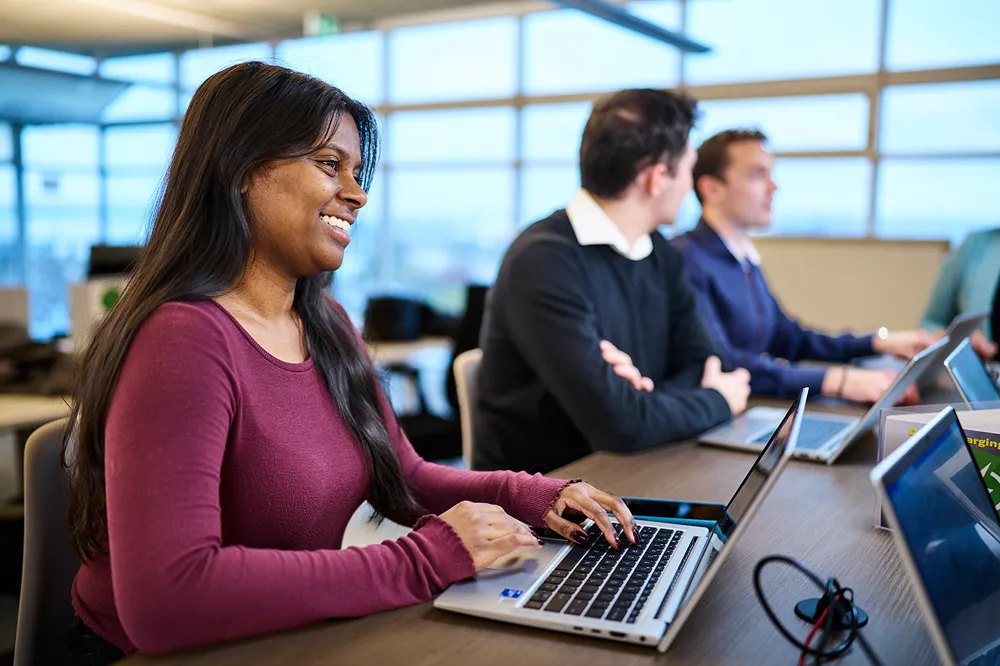 A smiling young professional is working on her laptop at a table with other colleagues