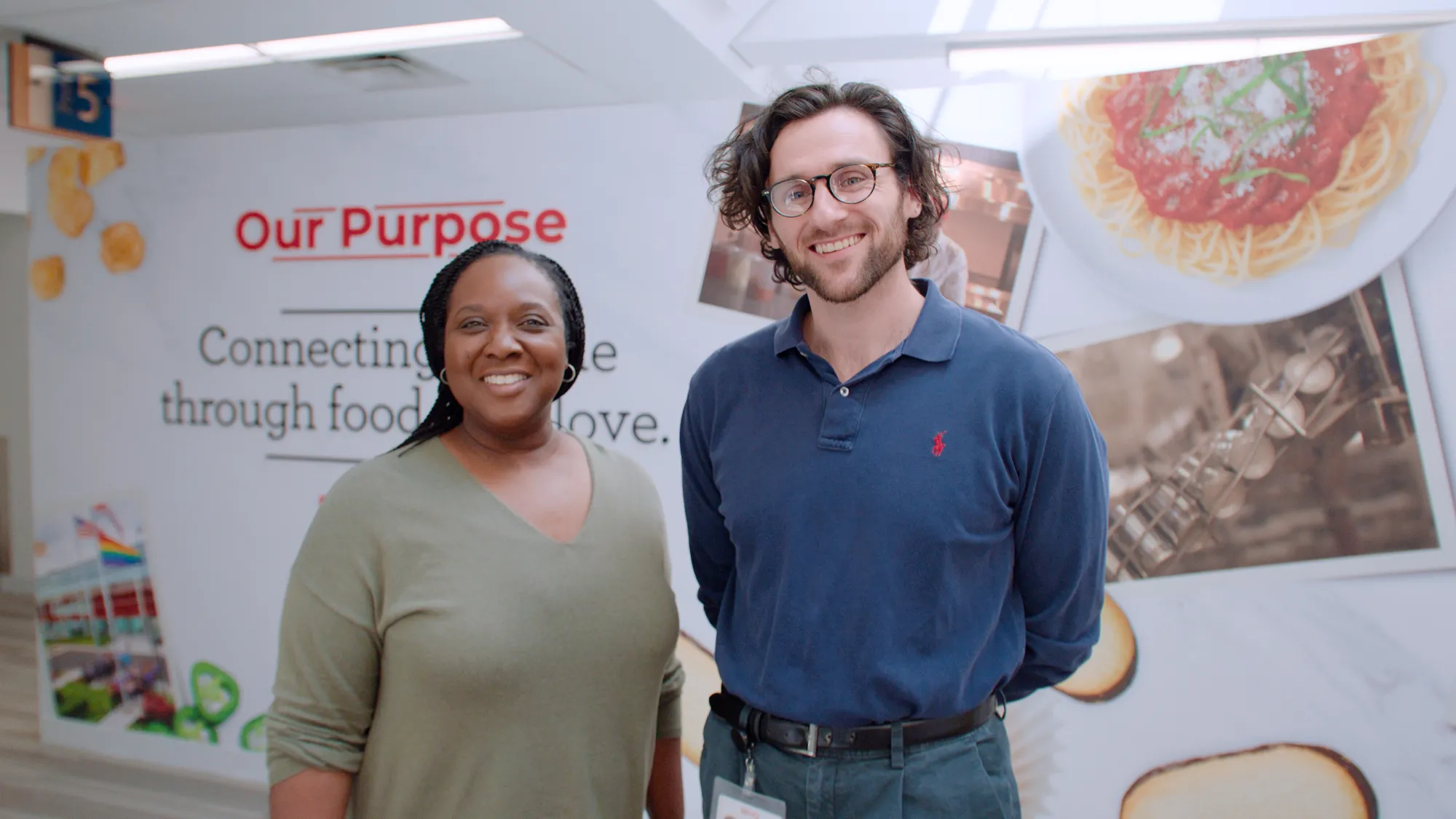 Two Campbell's employees smile for a photo in front of a wall showcasing the company's purpose: connecting through food and love