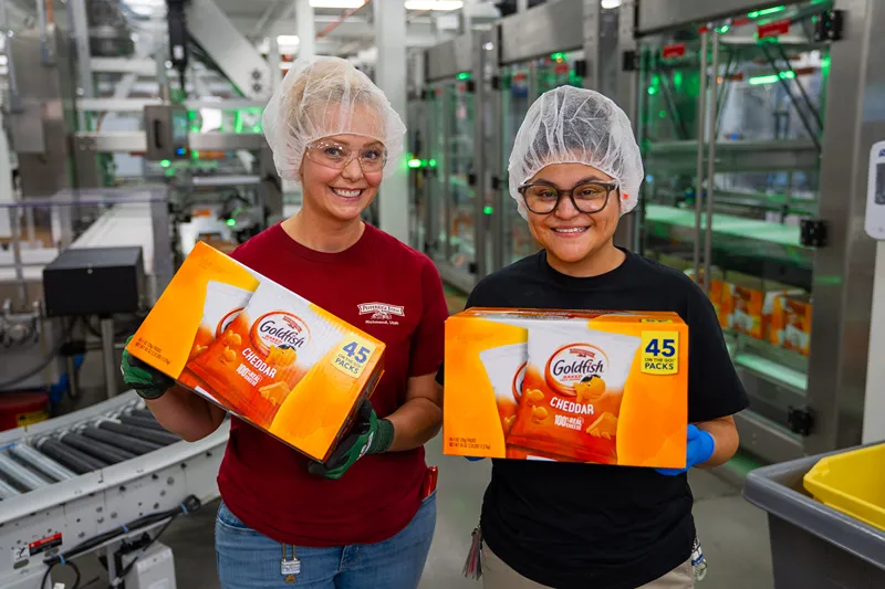 Two smiling female workers in a manufacturing plant hold boxes of Goldfish Cheddar crackers