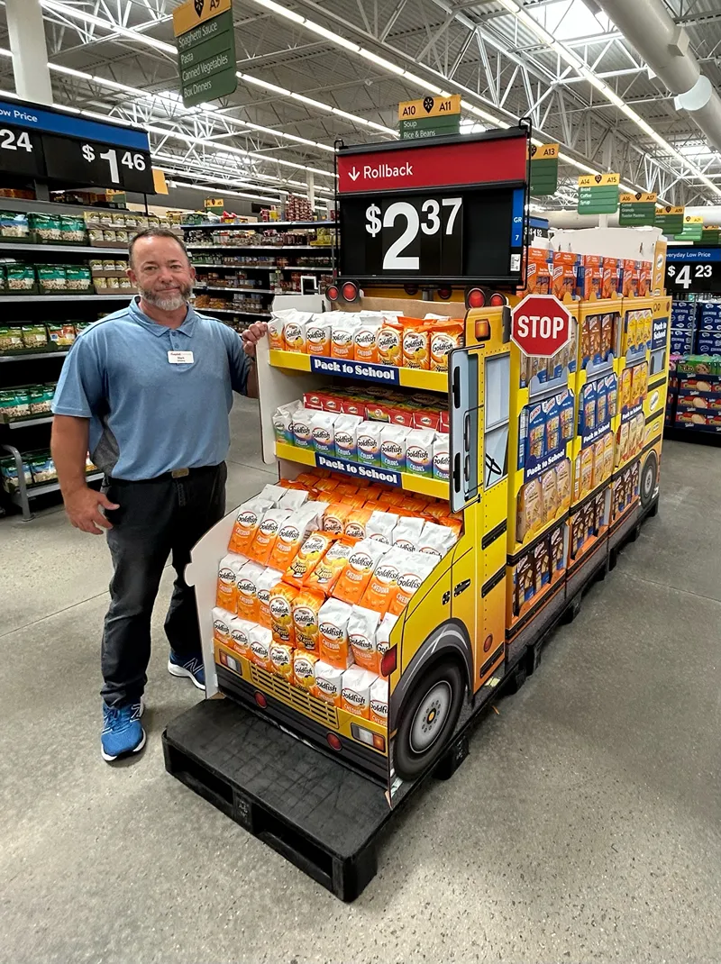 Walmart employee standing beside a school bus-shaped display of Goldfish crackers on sale for $2.37