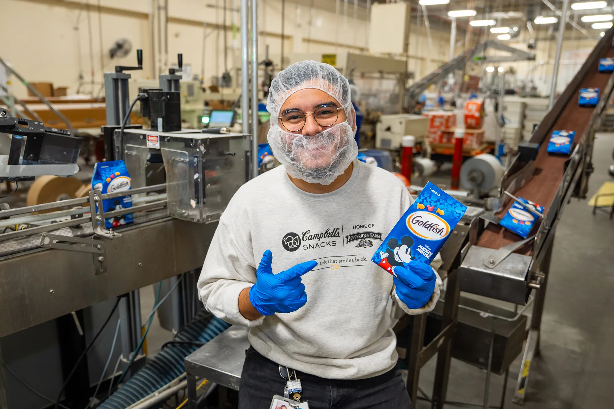 A smiling Campbell's employee in a factory setting holds a package of Disney Mickey Mouse Goldfish crackers while pointing towards the production line