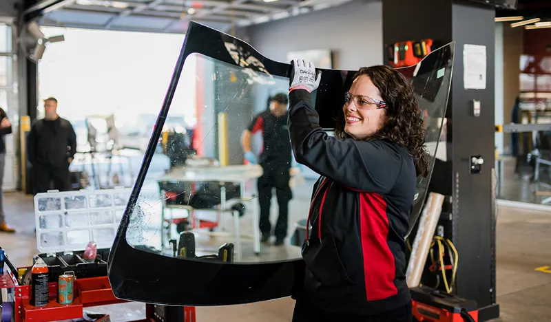 A smiling female technician in safety glasses and gloves holds a broken car windshield in an auto glass repair shop