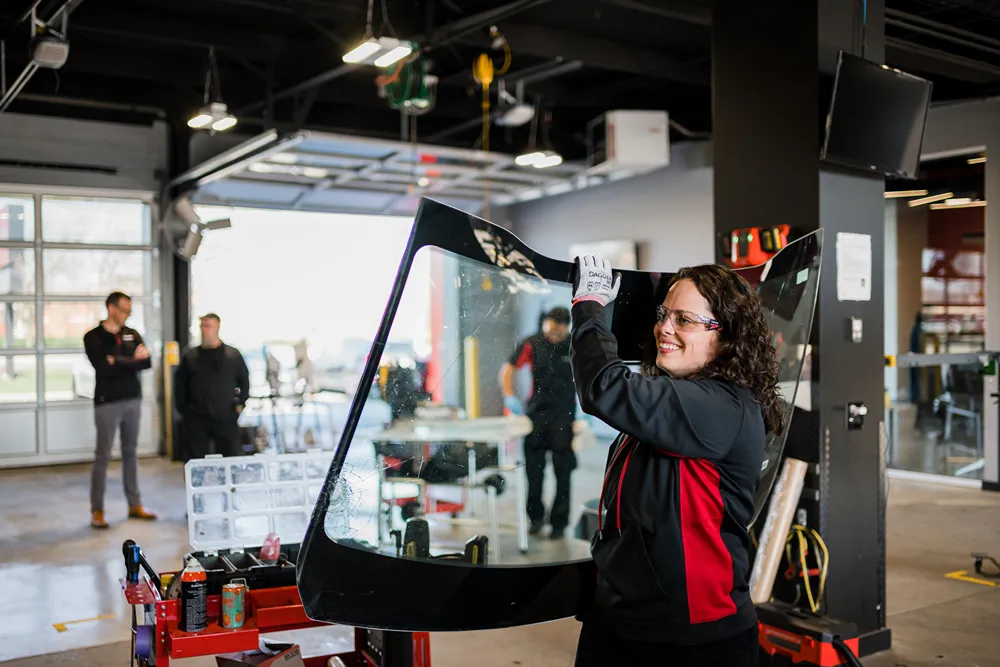 A smiling female technician in safety glasses and gloves holds a broken car windshield in an auto glass repair shop