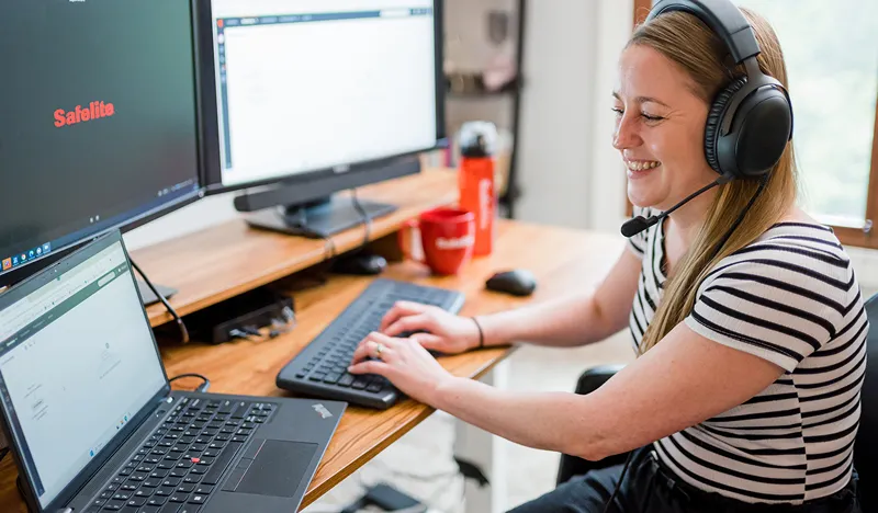 A smiling woman wearing a headset and striped shirt works at a desk with multiple computer screens, labeled 'Safelite'
