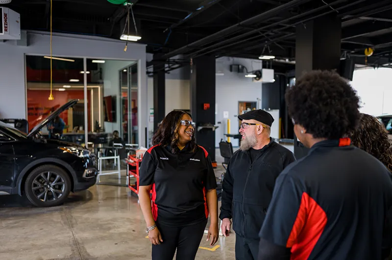 A group of Safelite technicians and managers discussing in a car repair shop, with vehicles in the background