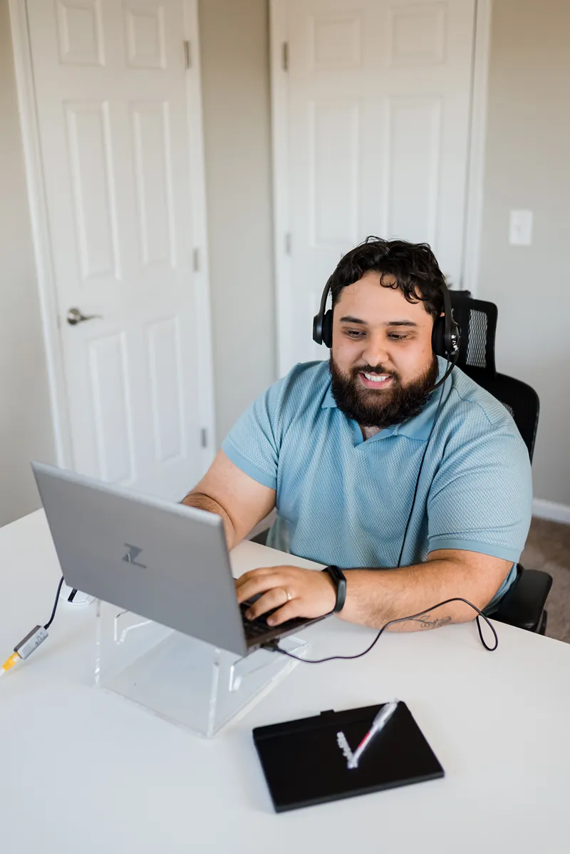 A smiling bearded man wearing a headset and blue polo shirt works on a laptop at a desk in a home office