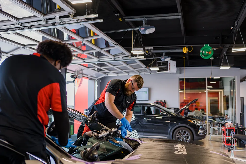 Two male Safelite technicians working on a car windshield replacement in a garage, one from the front and one from the back