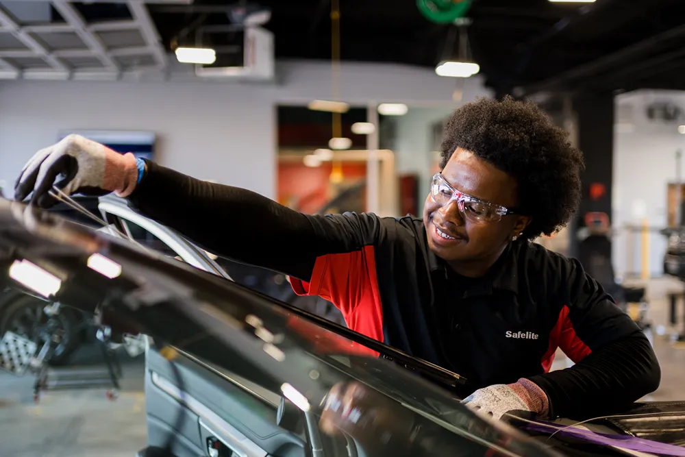 A smiling male Safelite technician in safety glasses and gloves, working on the side of a car