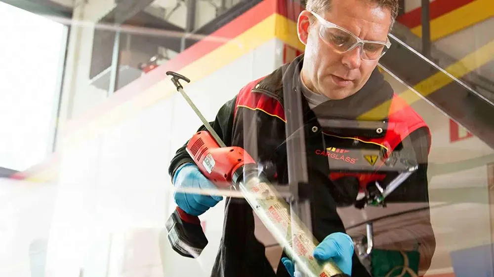 A technician in safety glasses and blue gloves applies sealant to a car window with a tool in an auto glass shop