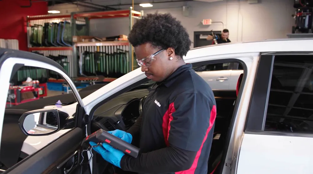 A male Safelite technician in safety glasses and blue gloves using a diagnostic tool inside a car.