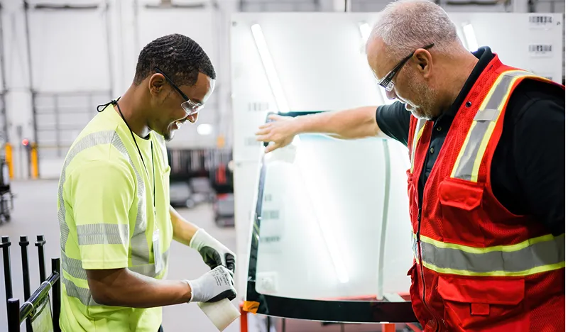 Two male workers in safety gear inspecting a large glass panel in a warehouse or factory