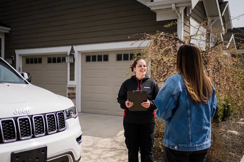 A Safelite technician with a clipboard speaking to a customer next to a white Jeep in a driveway