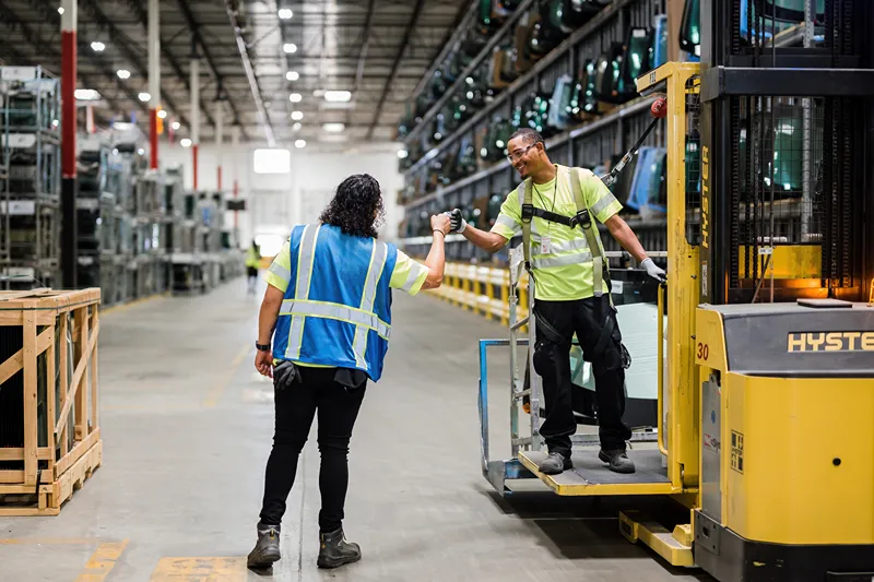 Two smiling warehouse workers in safety vests giving each other a fist bump near a forklift and rows of glass