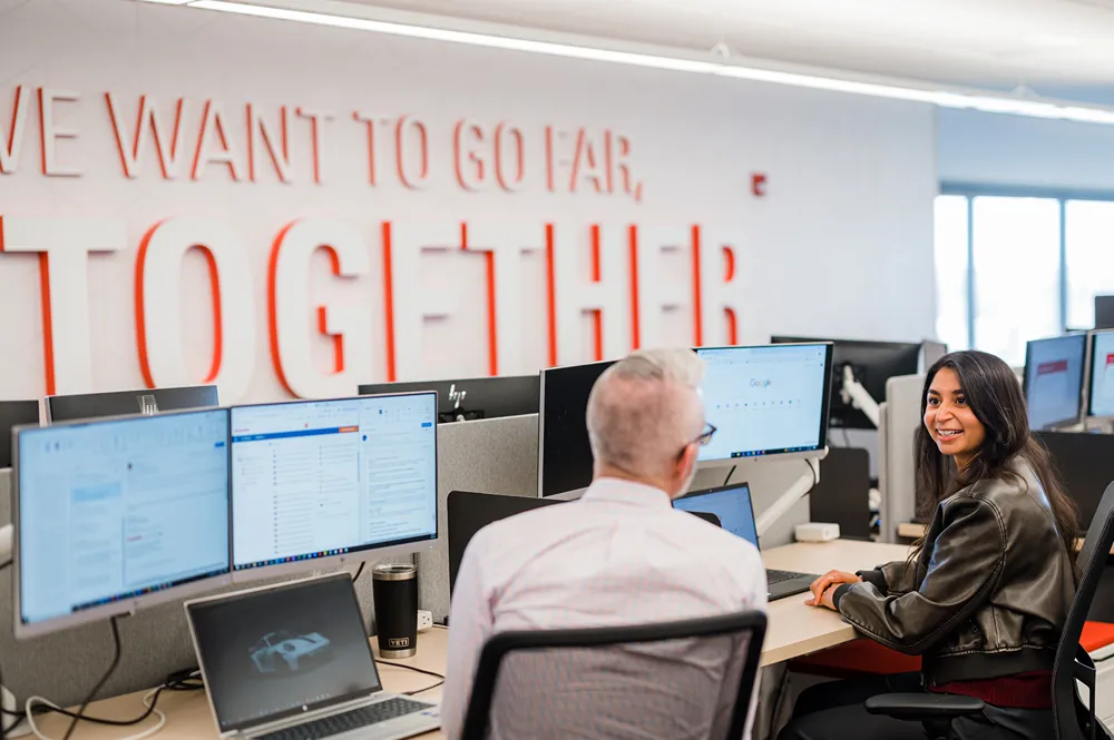 A young woman smiles at the camera while working on her computer at a desk in a finance office with 'We want to go far, together' on the wall