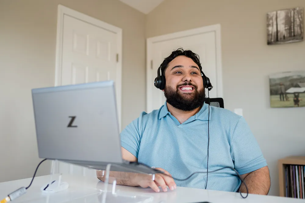 A smiling bearded man wearing a headset works happily on a laptop at a home office desk.