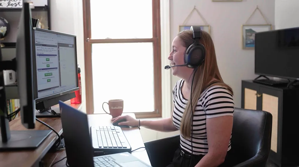 A woman wearing a headset and smiling while working at a computer in a home office