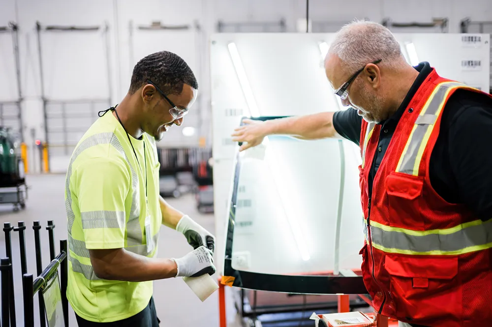 Two male warehouse workers in safety gear inspecting a large car windshield.