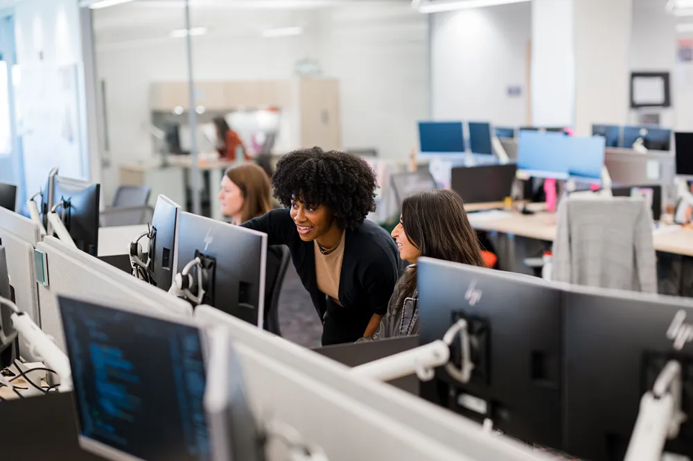 Two women collaborating and smiling at a computer workstation in a bustling open-plan office