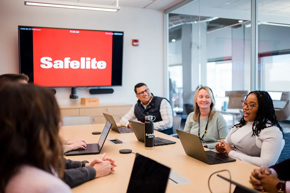 Diverse team meeting in a conference room with a large screen displaying the Safelite logo and a 'Safelite NEXT' water bottle on the table.