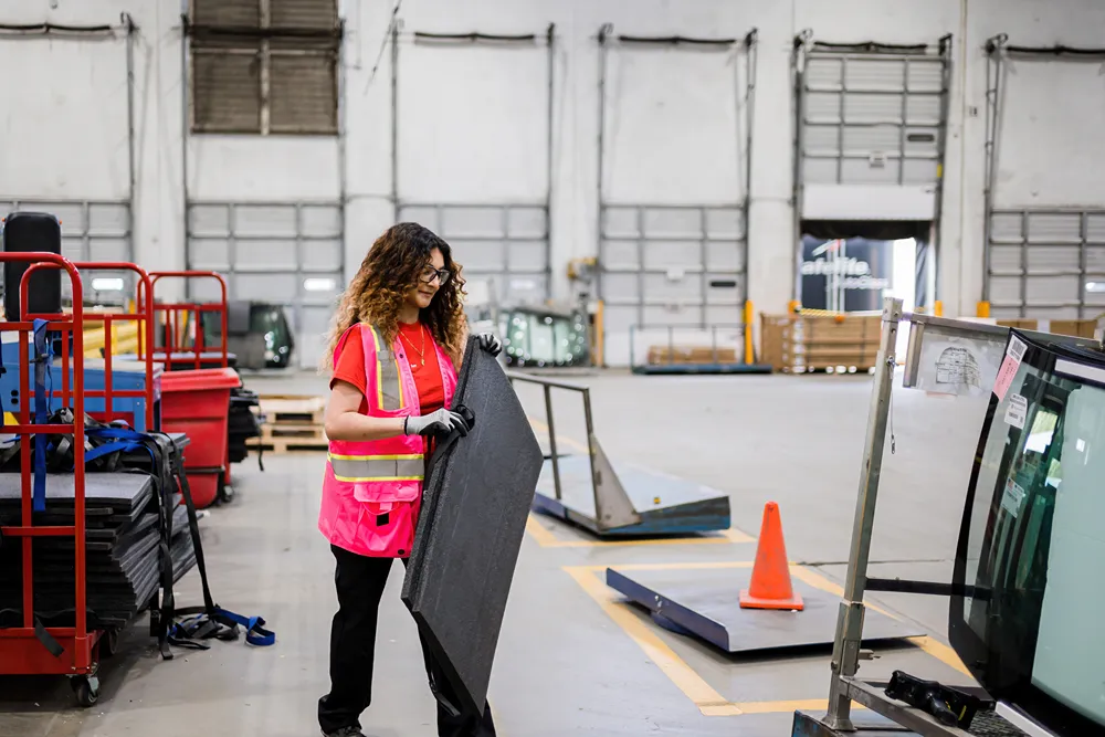 A female warehouse worker in a pink safety vest and gloves carrying a car part in a large distribution center.