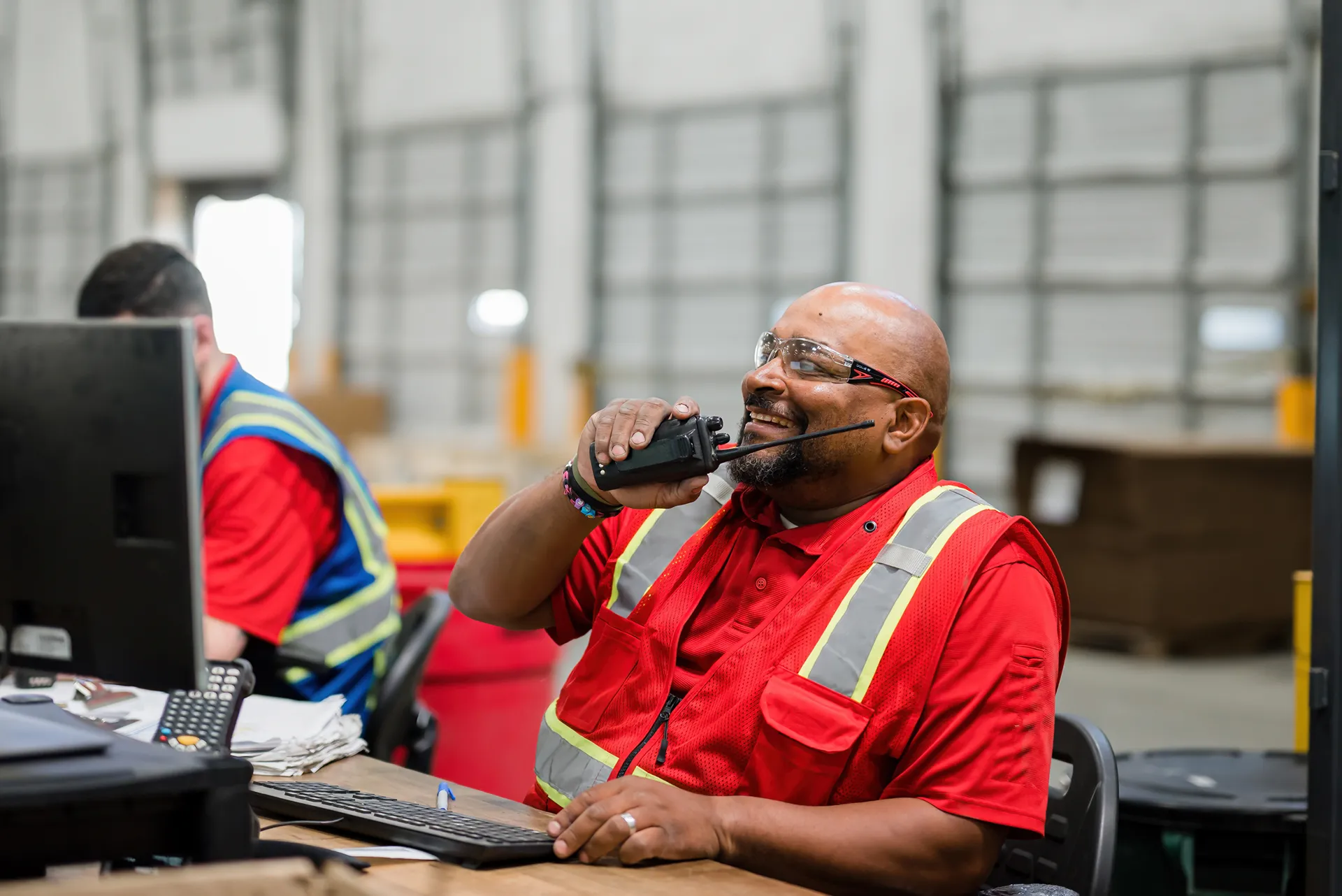 A smiling male warehouse worker in a red safety vest speaking into a walkie-talkie at a desk.