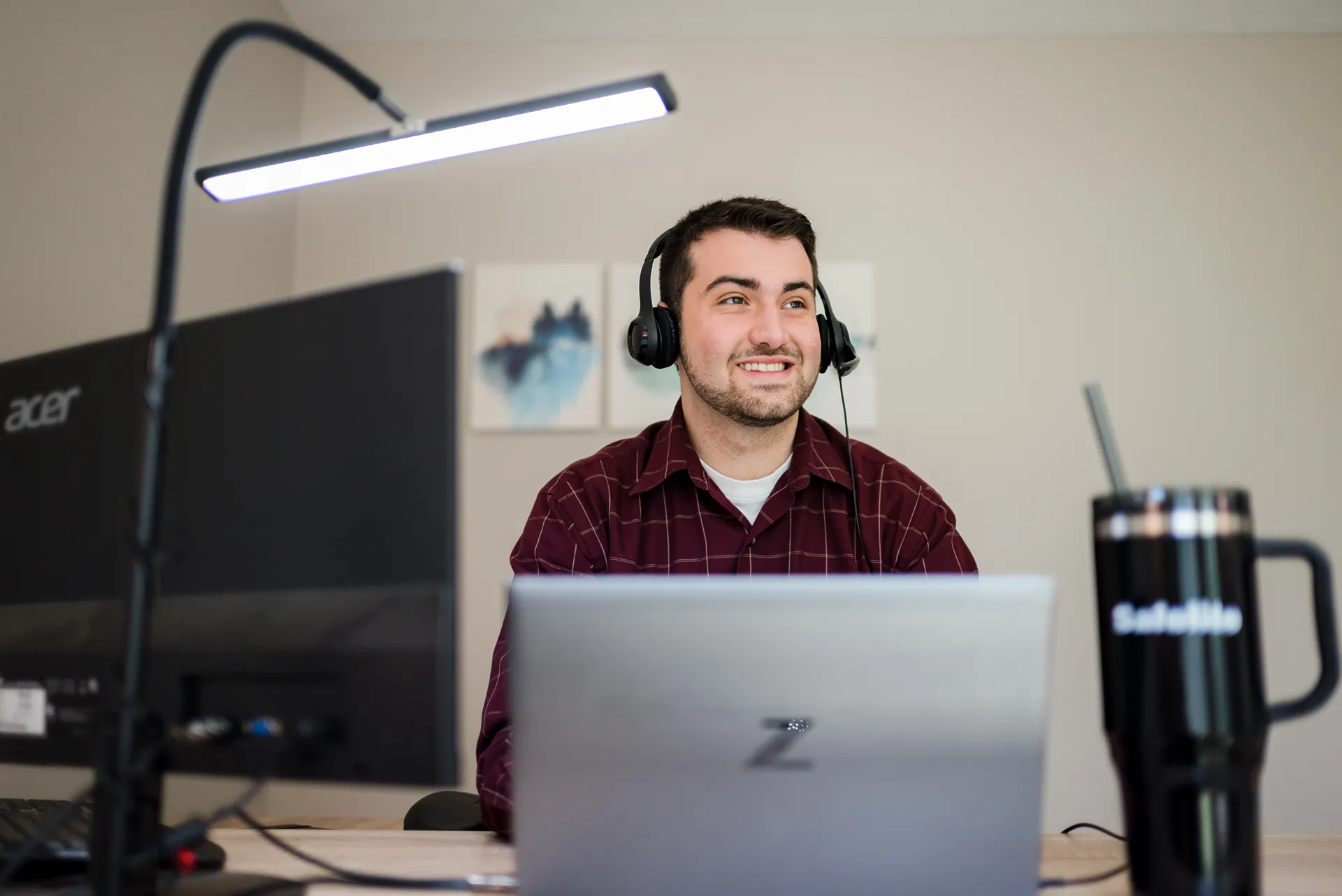 A smiling man wearing a headset and a plaid shirt works on a laptop and computer at a home office desk.