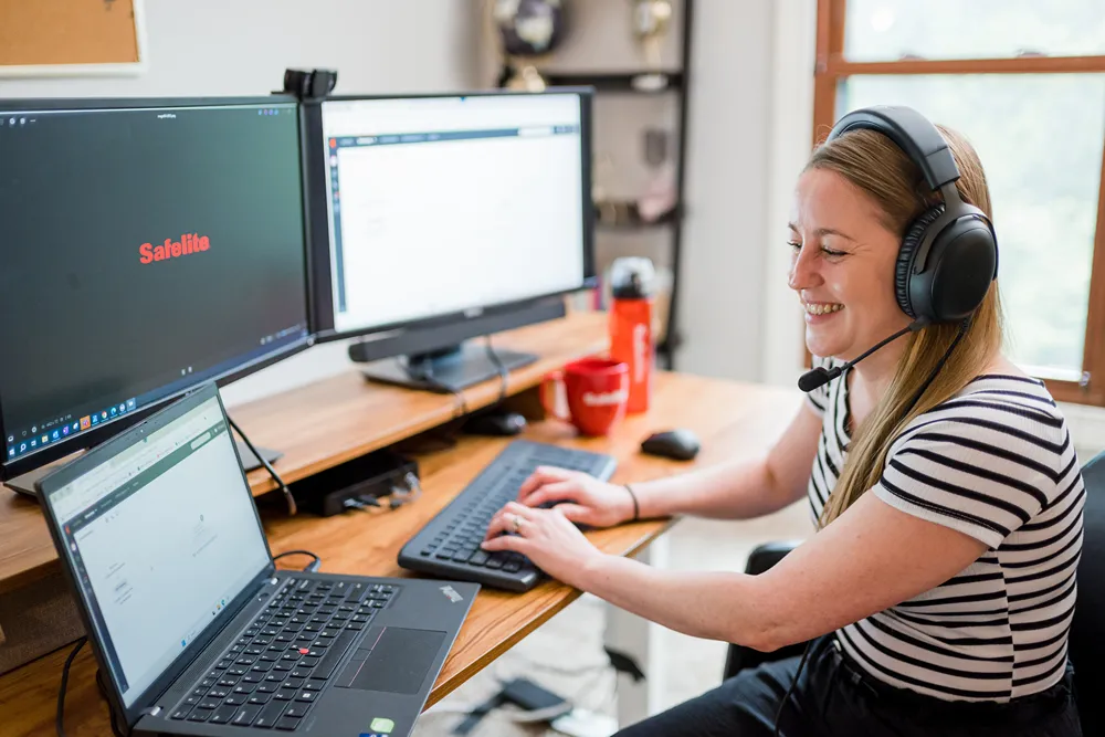 A smiling female Safelite customer service representative with a headset working at a home office desk with multiple monitors