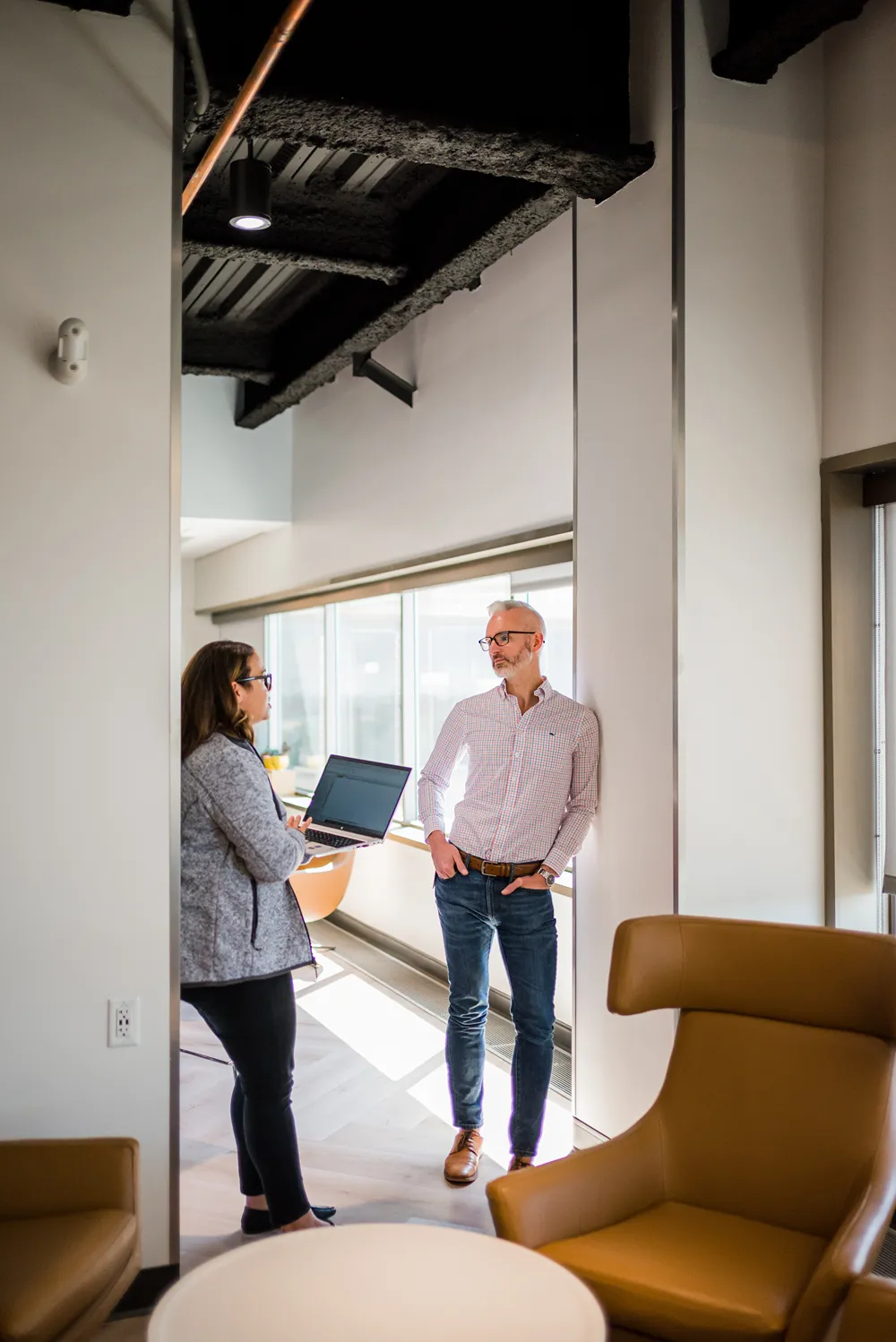 A woman holding a laptop stands and talks to a man in a modern, well-lit office hallway with large mirrors