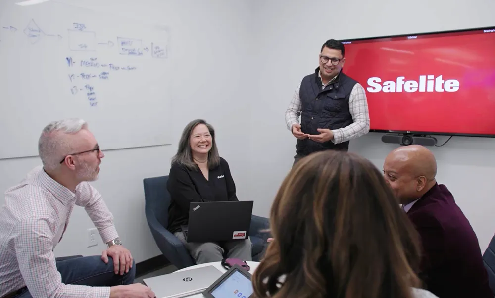 Diverse group of smiling professionals having a meeting in a conference room with a whiteboard and a screen displaying 'Safelite.'