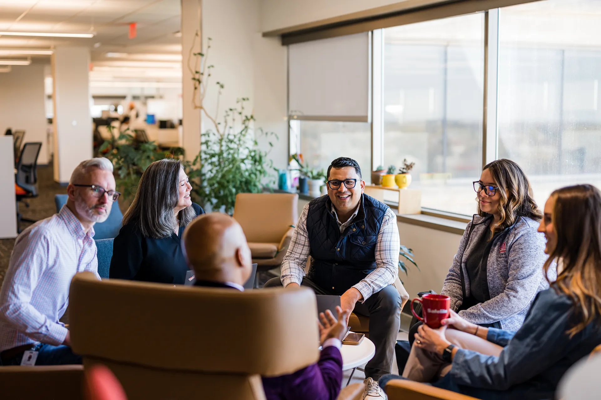 Diverse group of Safelite professionals laughing and talking in a casual meeting area with armchairs and plants.