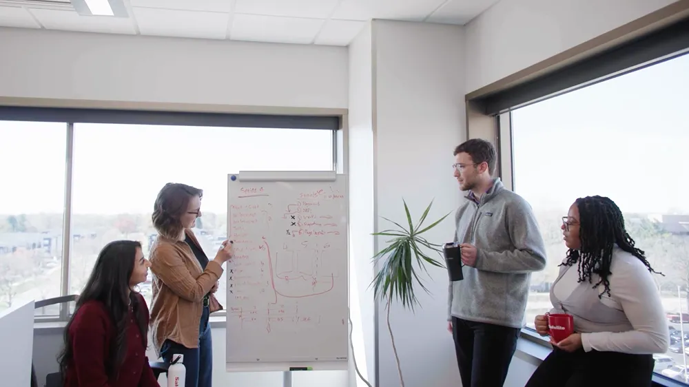 Diverse group of young professionals and a mentor discussing ideas around a whiteboard in a bright office.