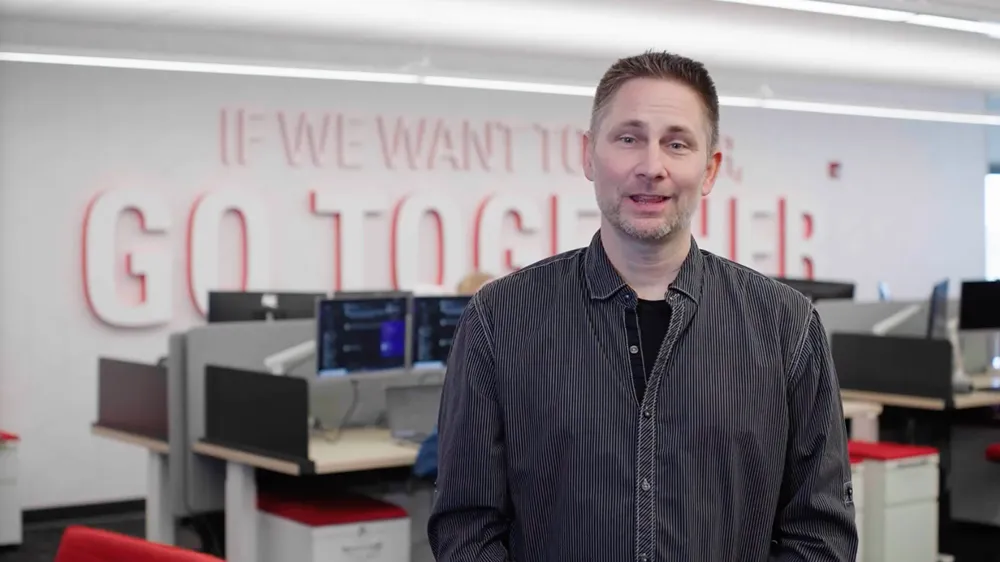A man in a striped button-down shirt smiles at the camera in a modern office with 'If We Want To Go Together' on the wall.