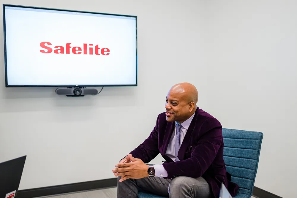 A smiling man in a purple jacket sits in a chair in a meeting room with a TV screen displaying the 'Safelite' logo