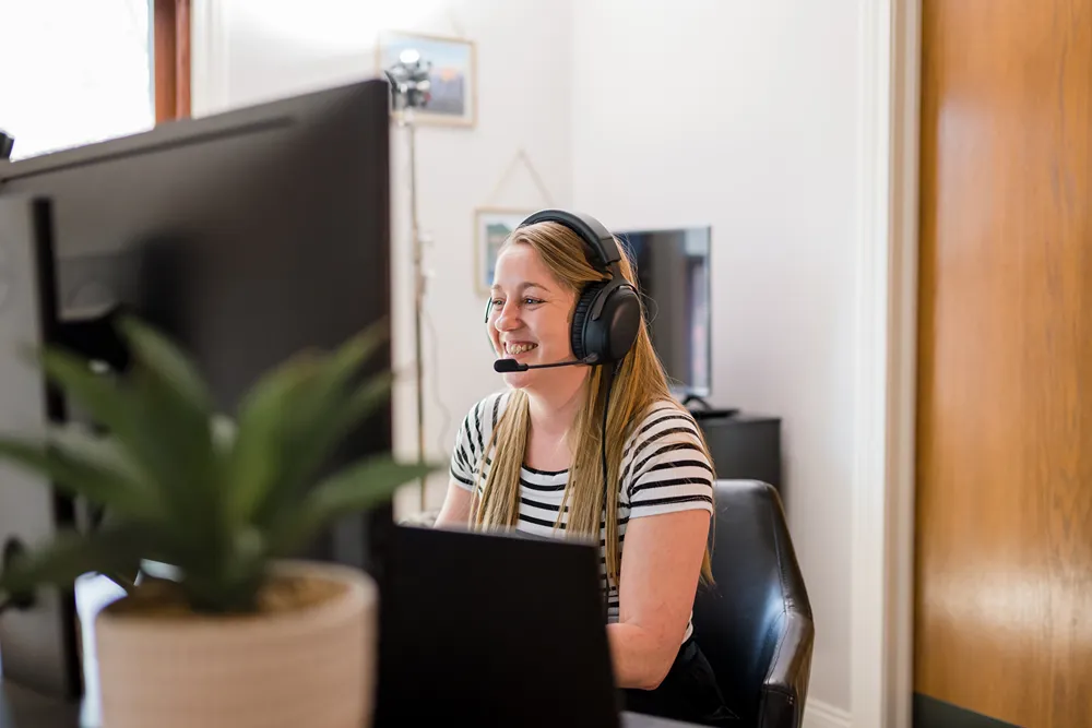 A smiling woman wearing a headset and striped shirt looks at a computer screen in a home office setting