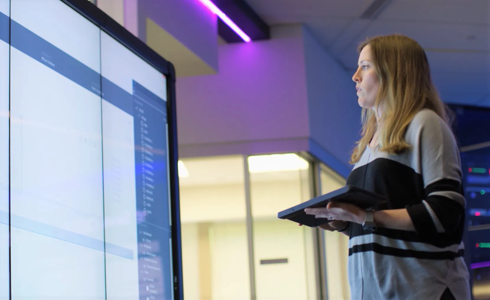 A woman holding a tablet in her hands looking at a large screen
