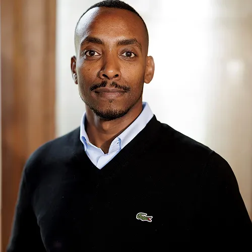 A headshot of a man wearing a black sweater and blue shirt looking intently at the camera