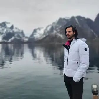 A photo of a man wearing a white coat standing next to a body of water and mountain can be seen in the background