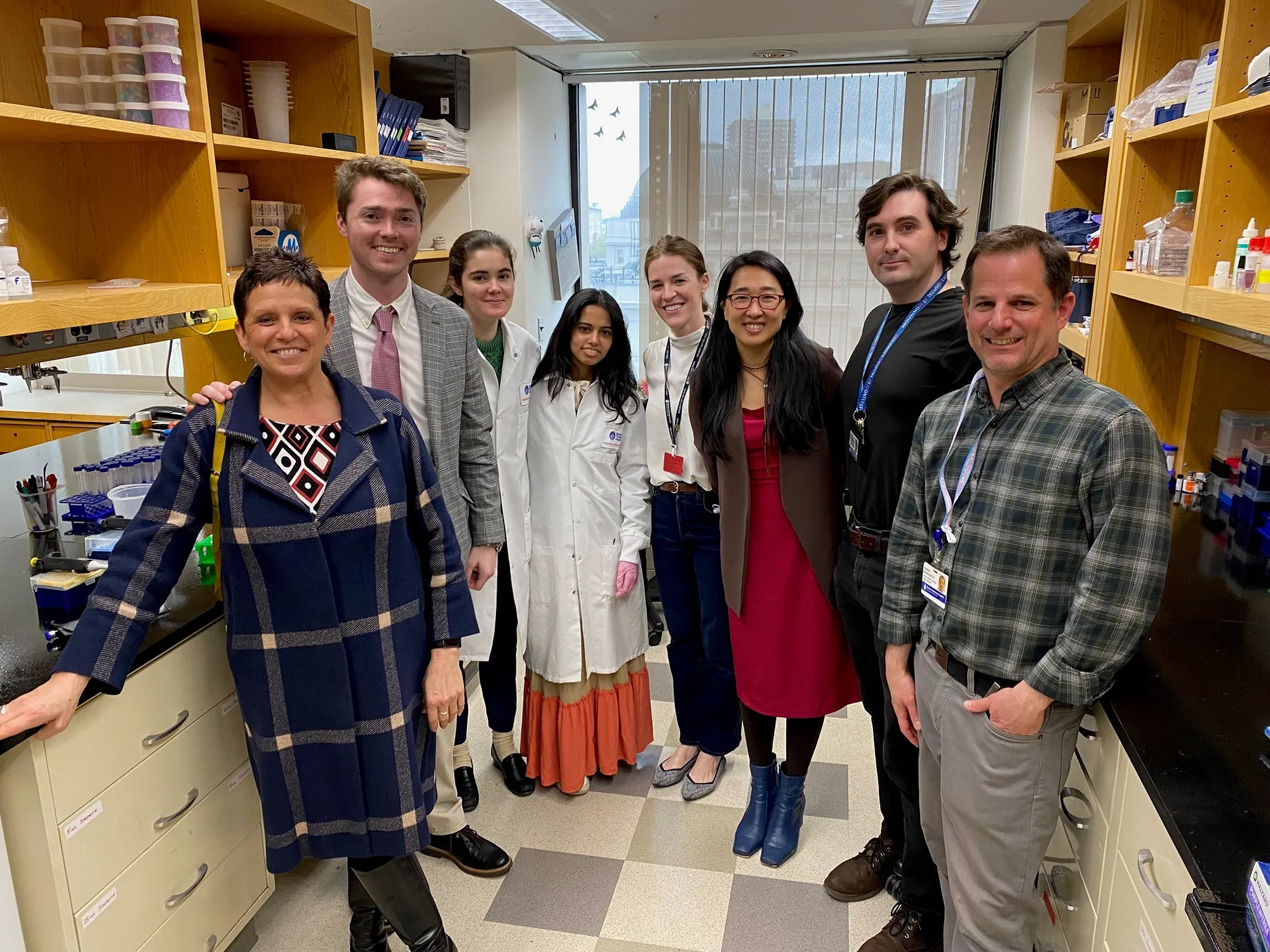 A group of eight people in a laboratory setting with shelves filled with lab supplies