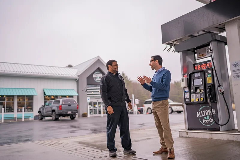 Two men talking near a gas station pump with a building in the background