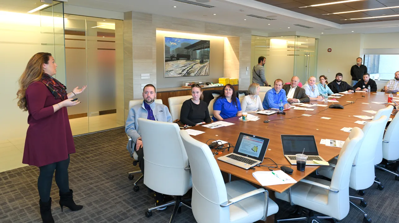 Business meeting in a modern conference room with a woman presenting to a group of professionals seated around a large table with laptops and documents.