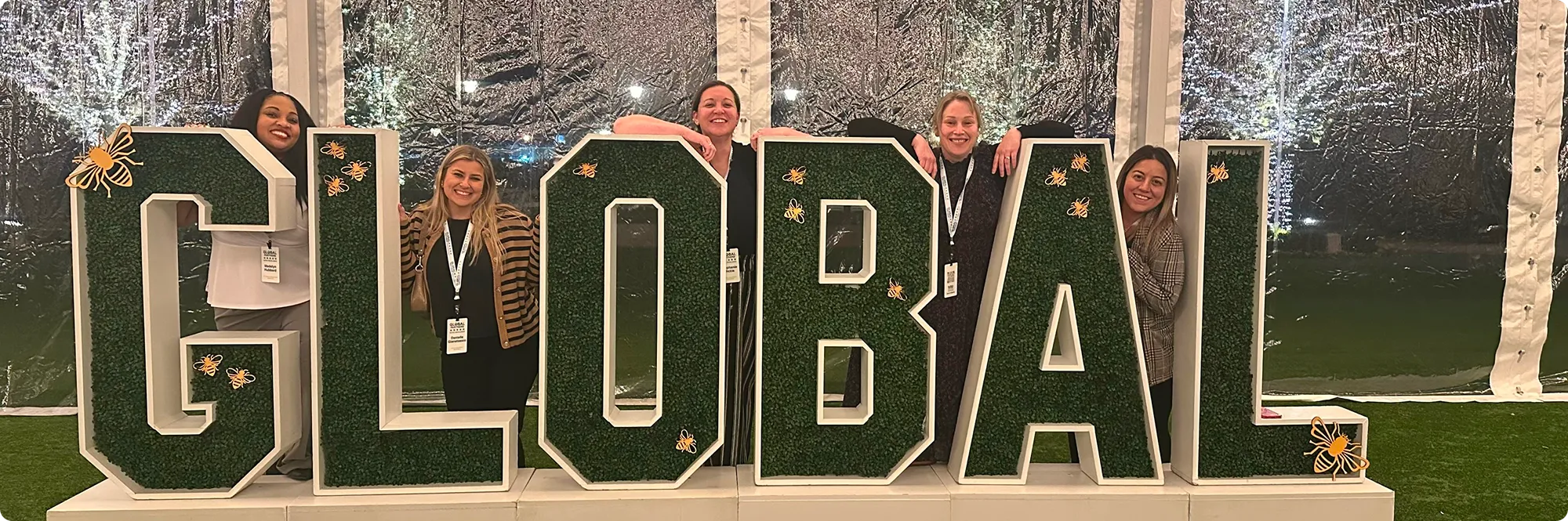 Group of smiling women posing behind large decorative letters spelling ‘GLOBAL’ at an event, symbolizing teamwork and international collaboration.