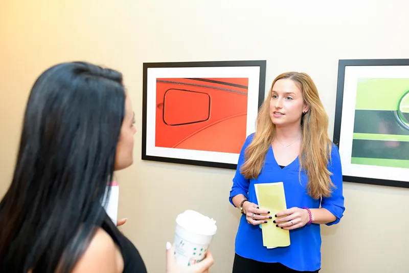 Two women talking in an art gallery, one holding a folder, the other holding a coffee cup, with abstract art in the background