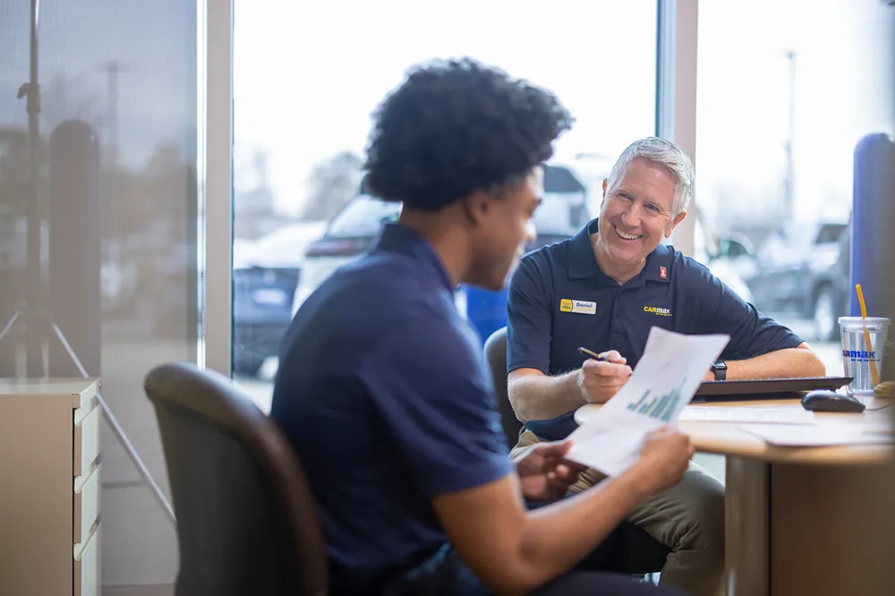 A smiling CarMax mentor is discussing documents with a young colleague in an office.